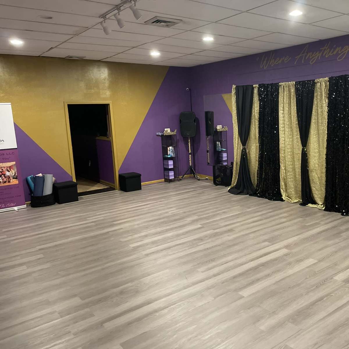 A spacious dance studio with a hardwood floor and a backdrop of black and gold sequined curtains against a purple wall.