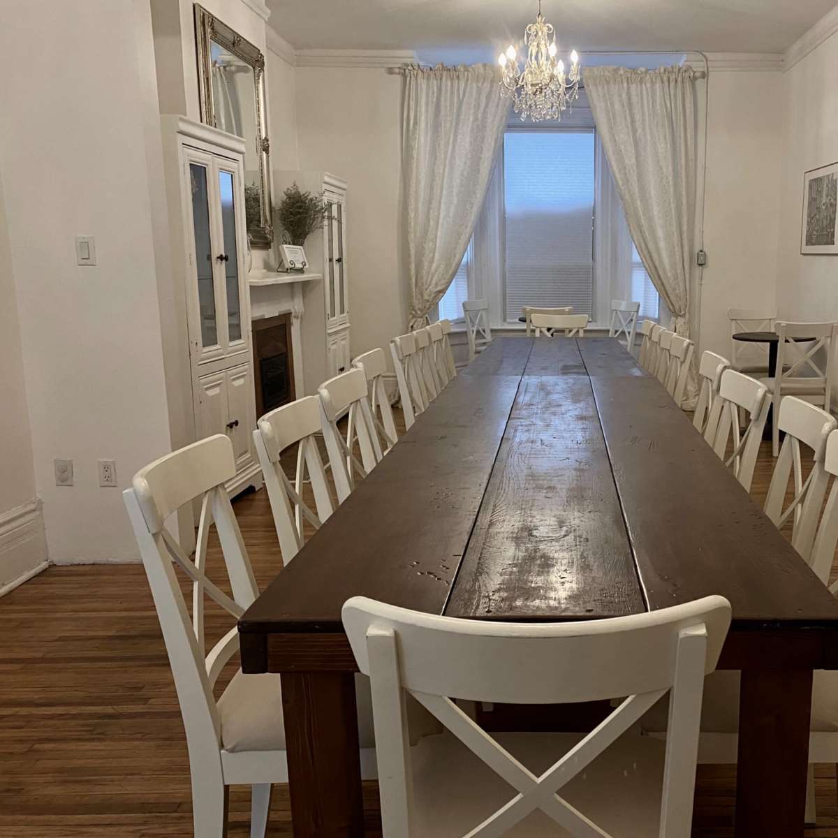 A long wooden dining table is lined with white chairs in a bright, spacious room featuring a chandelier and large windows.