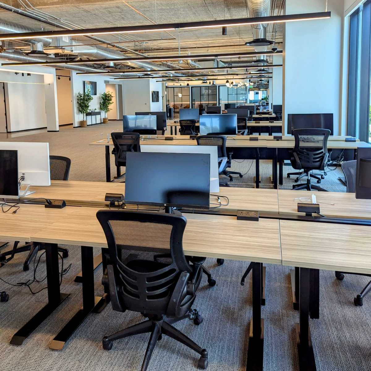 A spacious, modern office with rows of desks, each equipped with computers and ergonomic chairs, and large windows letting in natural light.