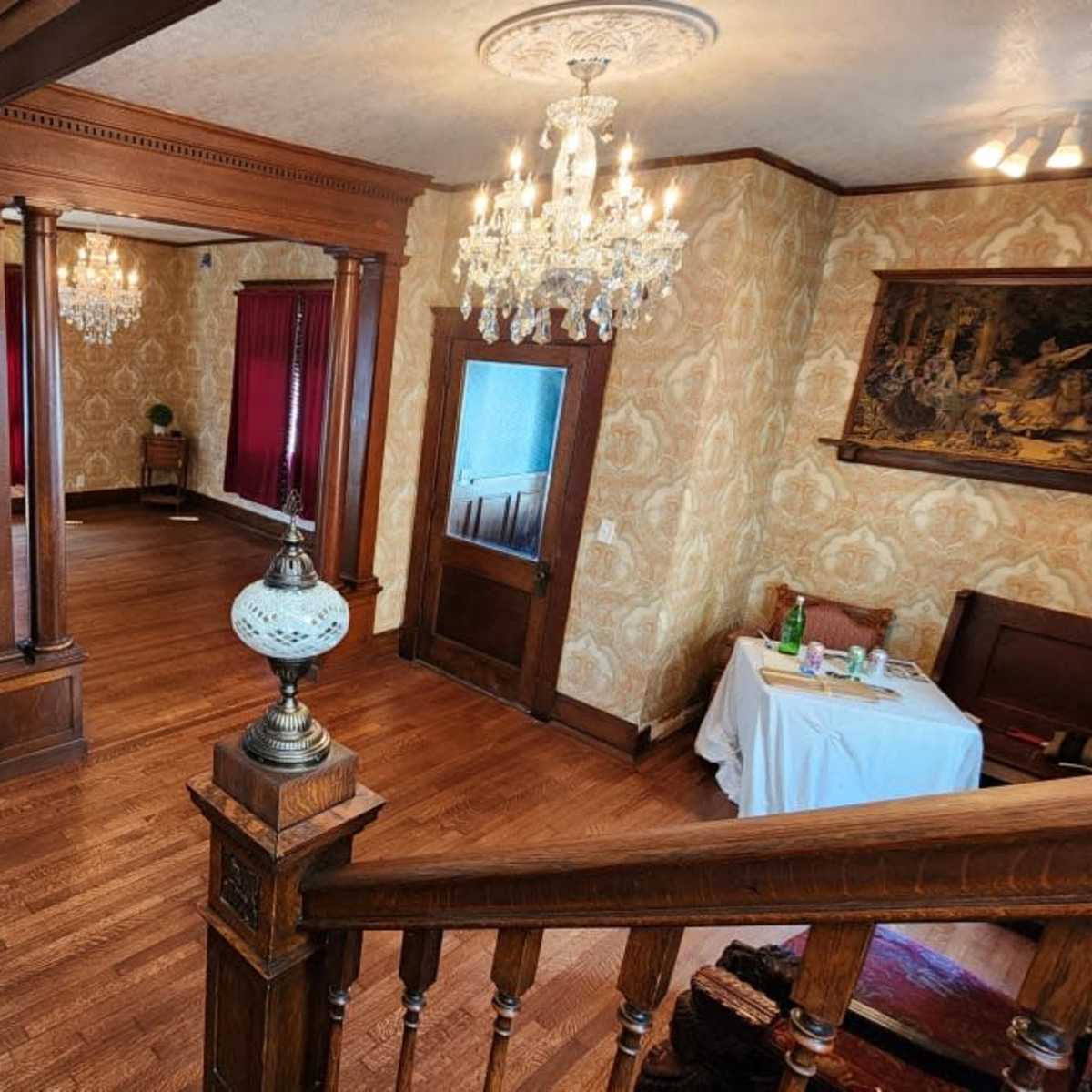 A spacious, vintage-style interior with hardwood floors, ornate wallpaper, and chandeliers, featuring a staircase leading to a well-furnished area with a table and chairs.