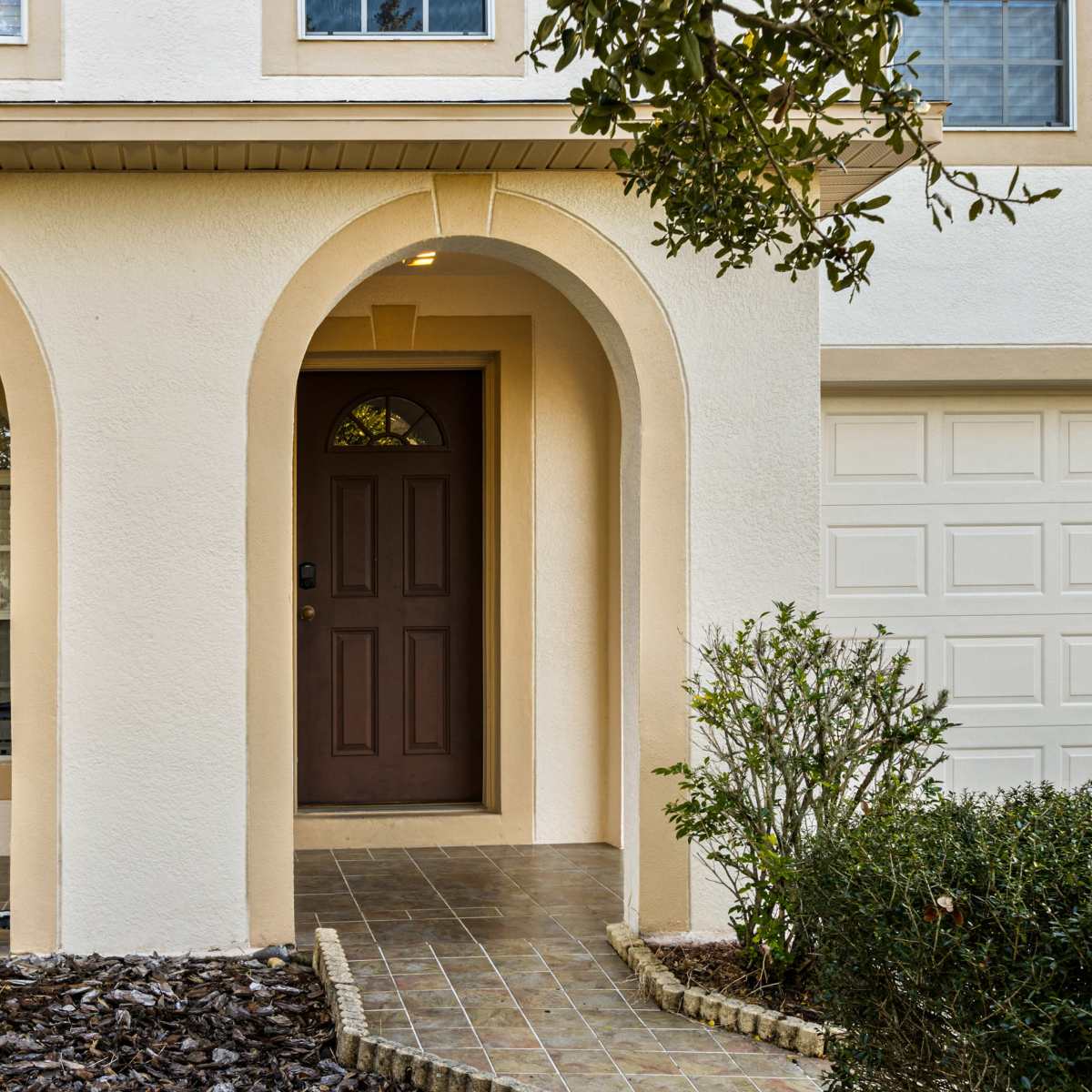 The entrance of a house with arched doorways, a brown front door, and a two-car garage visible in the background.