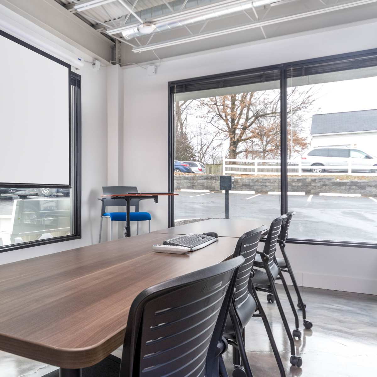 A conference room features a large table, black chairs, a projector screen, and large windows overlooking a parking lot.
