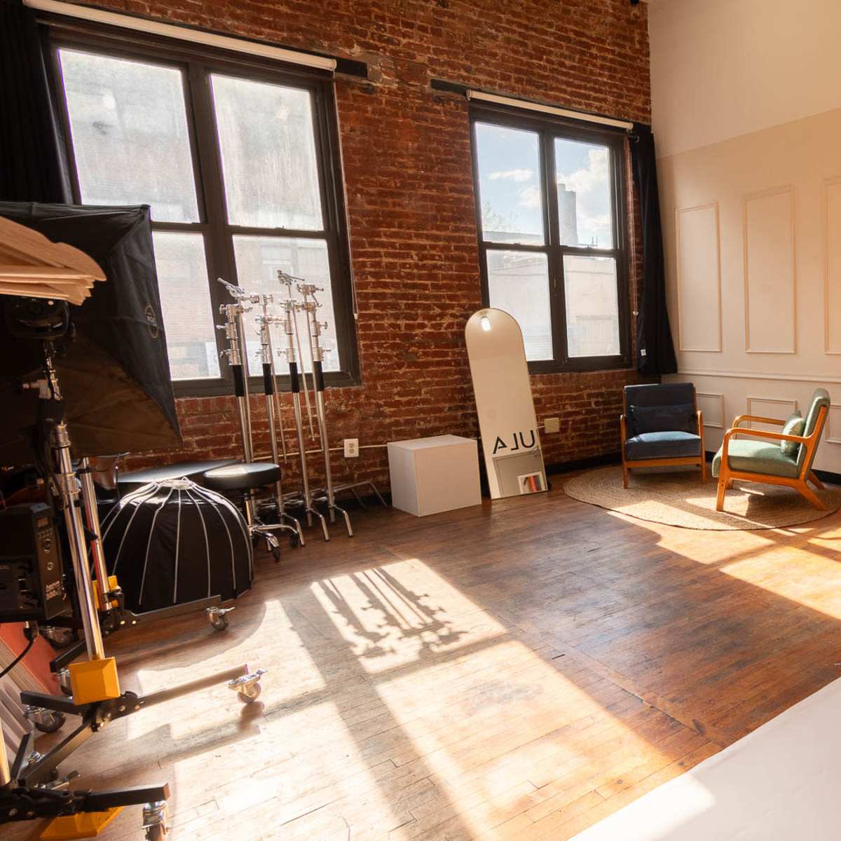The room features a wooden floor, a stool in the center, and a wall with colorful paper rolls alongside a vintage chair and a large window letting in natural light.