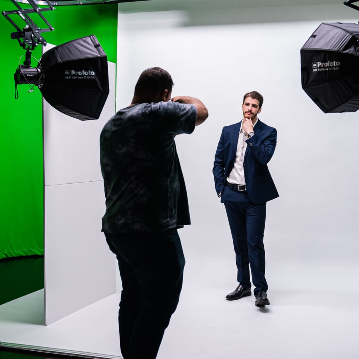 A photographer is taking a picture of a man in a suit against a plain white backdrop illuminated by studio lights in a green screen environment.