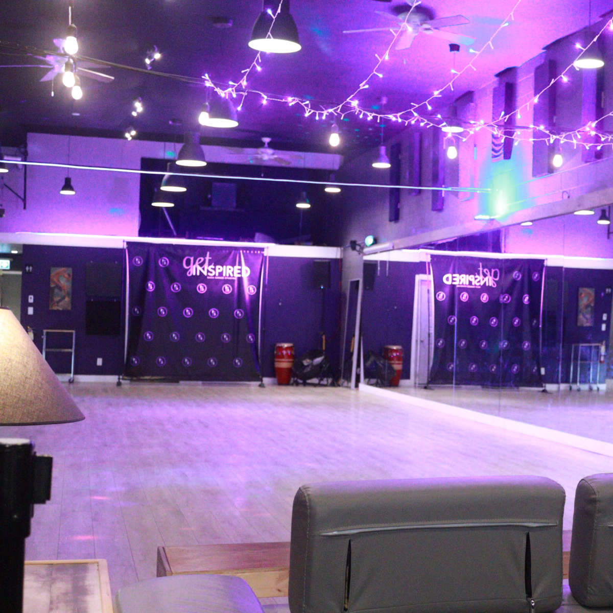 A dance studio with wooden flooring, mirrored walls, and purple lighting, featuring decorative string lights overhead.