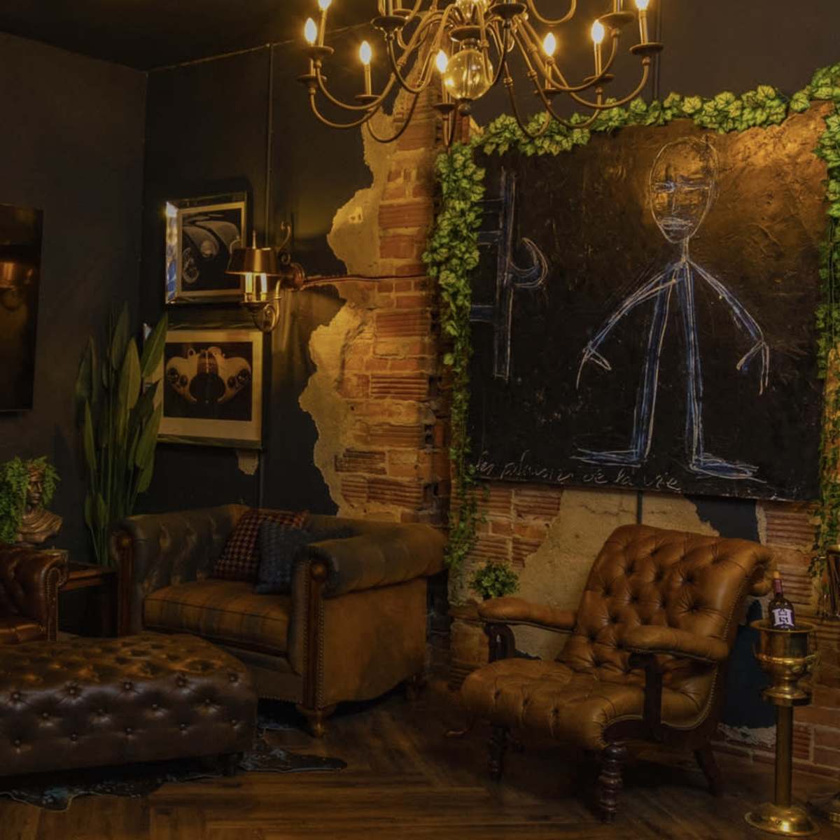 A cozy lounge area featuring a dark wall, a large flat-screen TV, a leather chesterfield sofa, an armchair, and a decorative blackboard surrounded by greenery.
