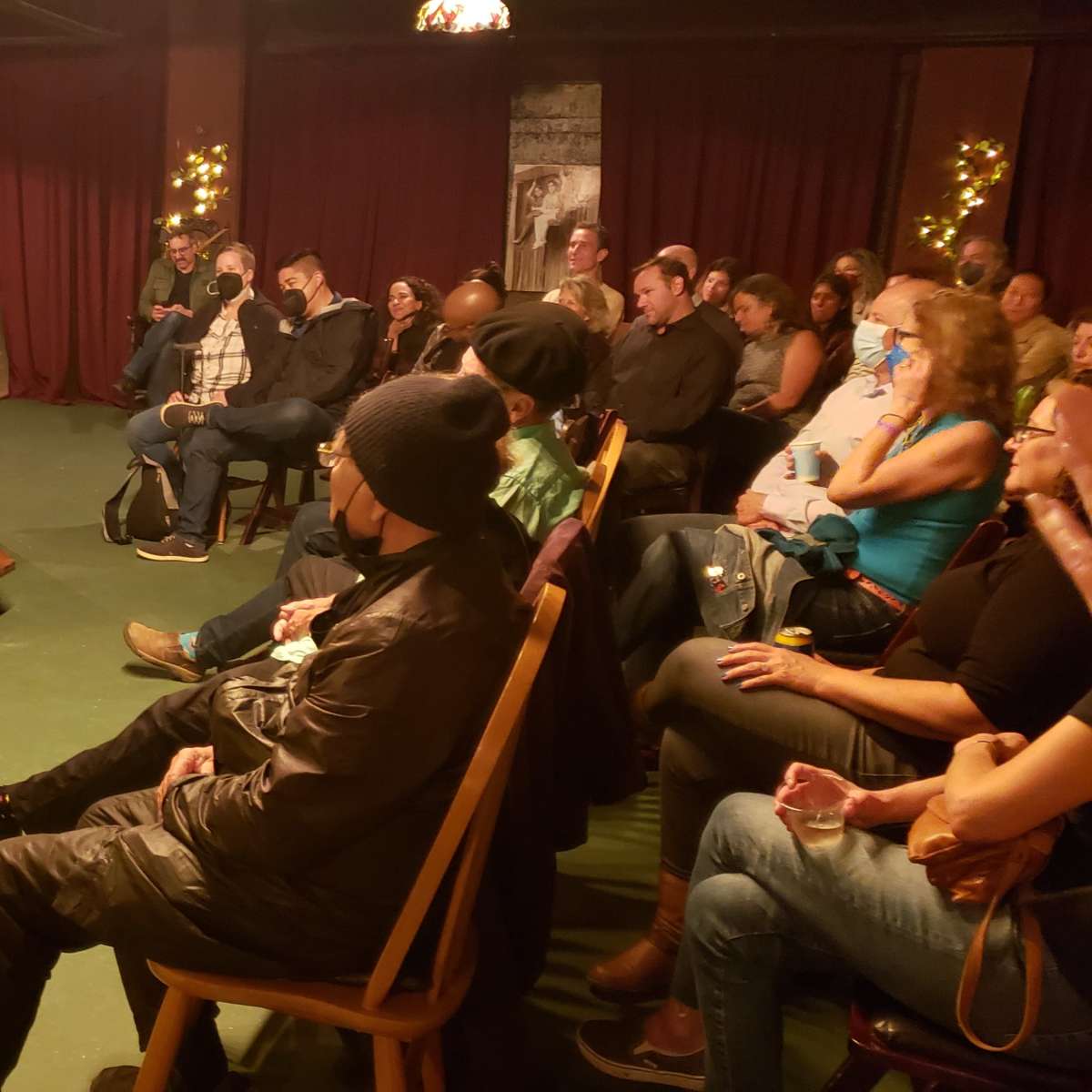A woman reads on stage while an audience sits attentively in a dimly lit venue decorated with lights and plants.