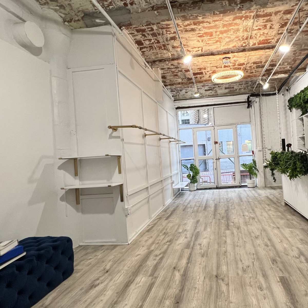A spacious interior with wooden flooring, a brick ceiling, and minimalist decor, featuring a blue upholstered bench and potted plants near glass doors.