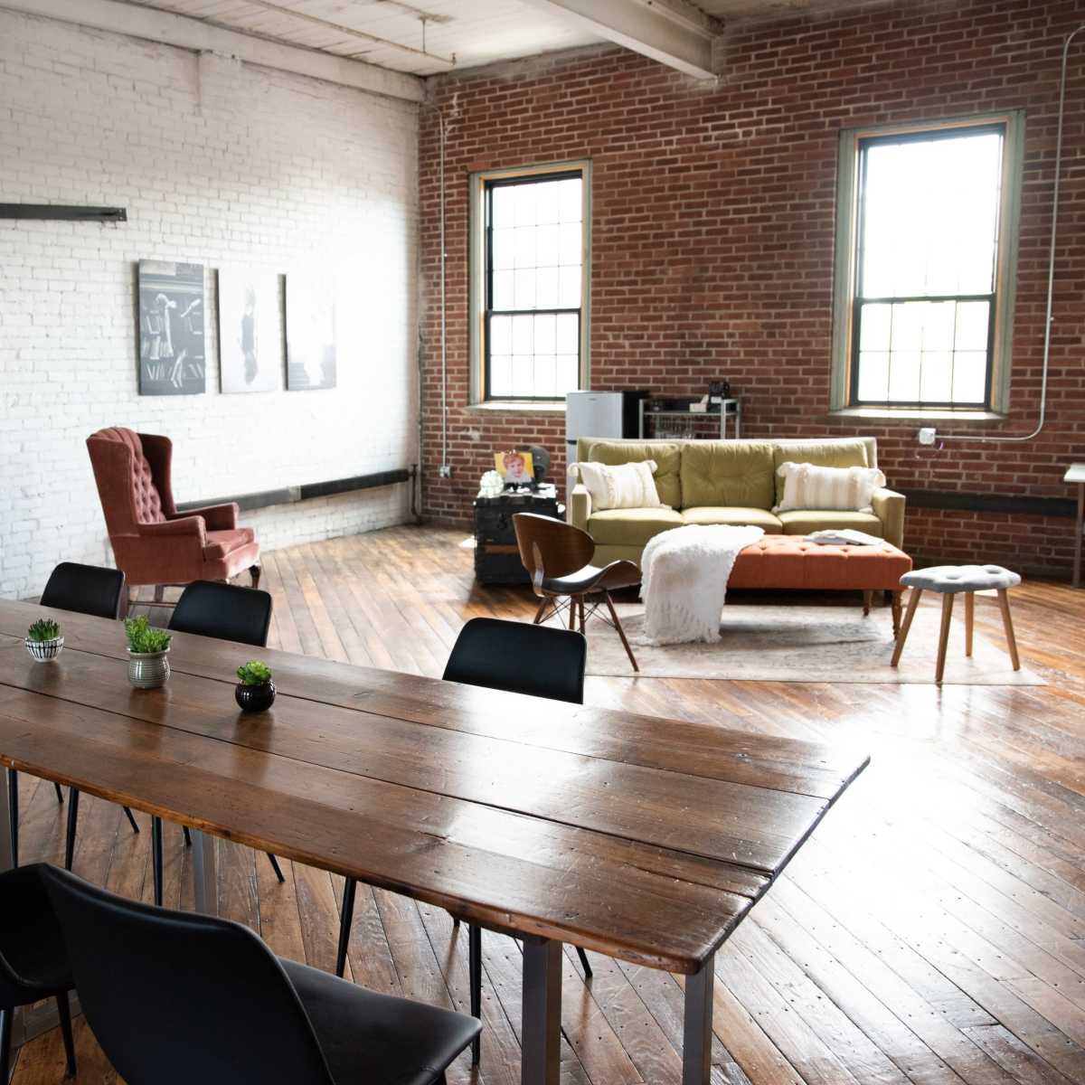 A spacious industrial room features a large wooden table with black chairs, a red accent wall, and a cozy seating area with a sofa and armchair.