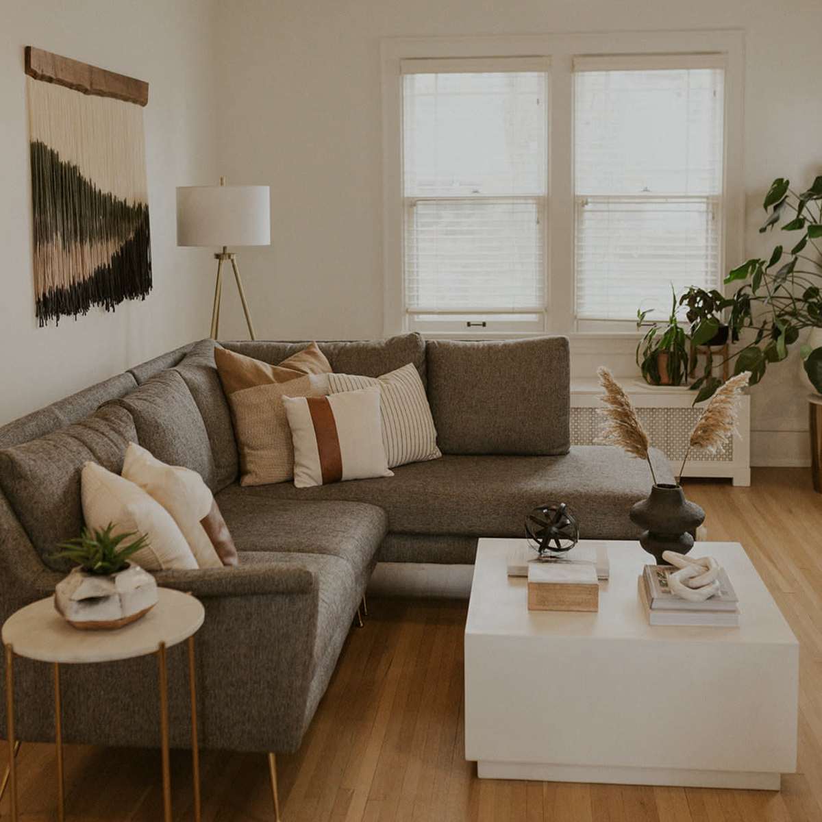 A modern living room featuring a gray sectional sofa with decorative pillows, a white coffee table with various decor items, and large windows allowing natural light in.