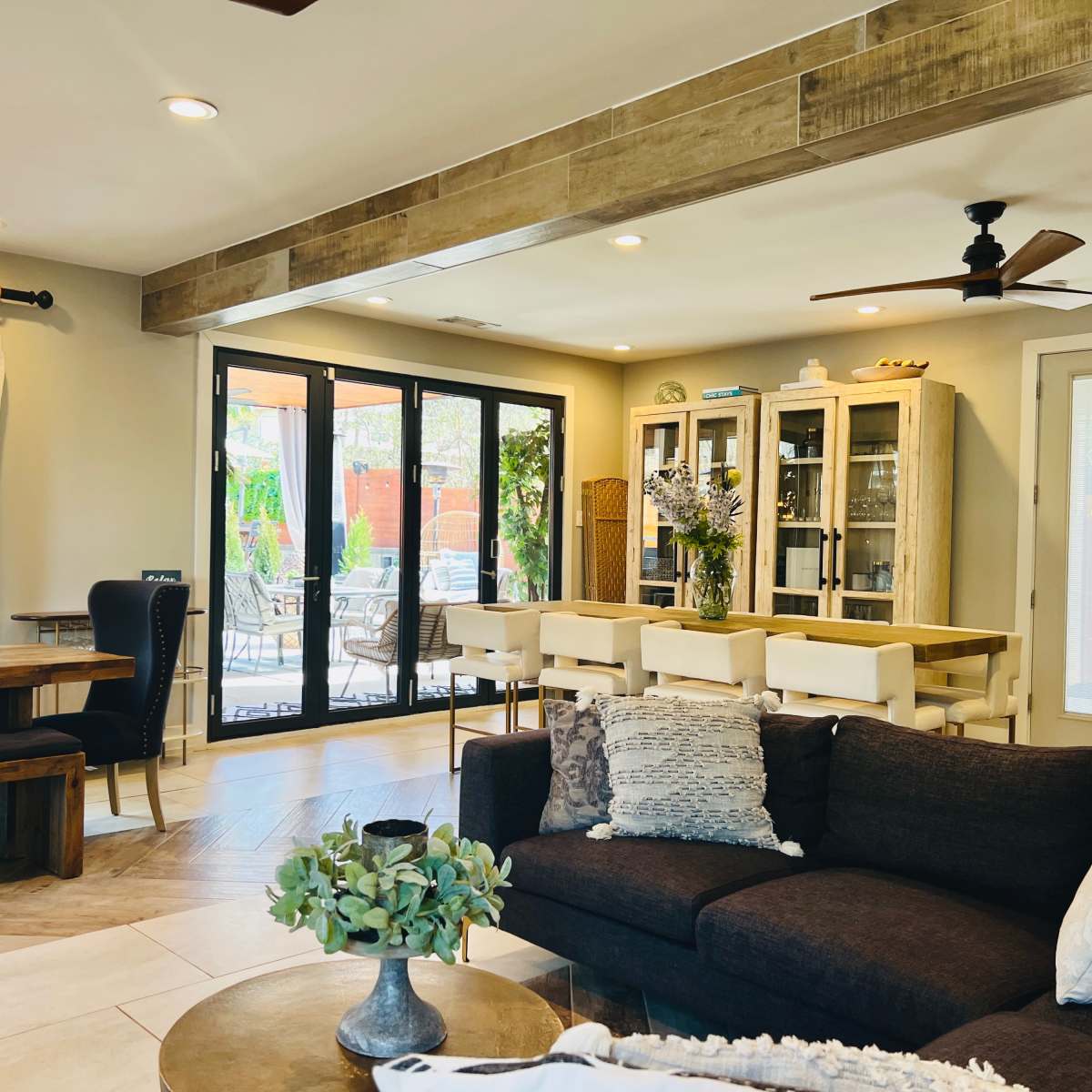 An open-concept living space featuring a dark sectional sofa, a wooden dining table, and large glass doors leading to an outdoor area filled with greenery.