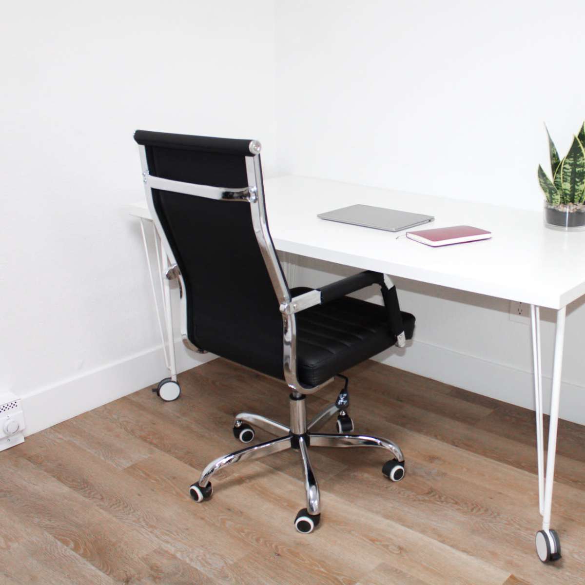 A minimalist office setup features a white desk, a black rolling chair, a laptop, a notebook, and a potted plant.