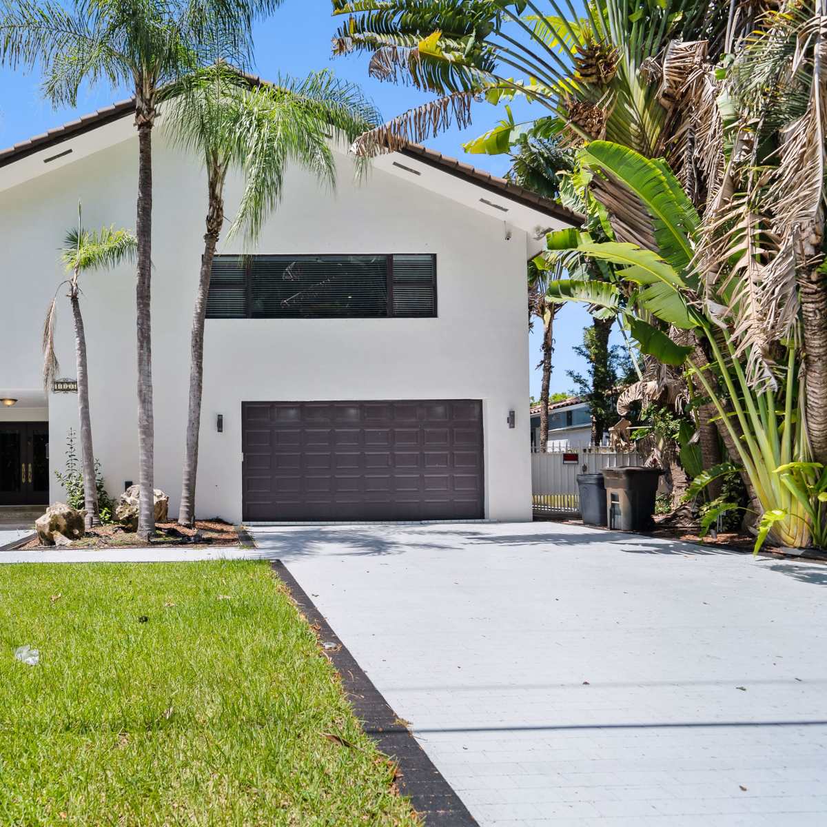 A modern two-story white house with a gray garage door, surrounded by palm trees and lush greenery.