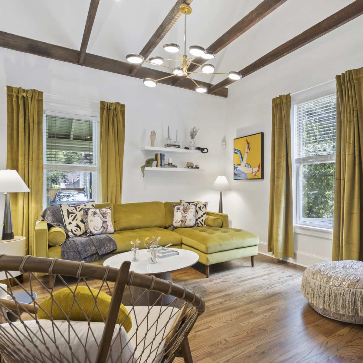 The living room features a green sectional sofa, a round coffee table, and two framed artworks on the walls, complemented by yellow curtains and wooden beams overhead.