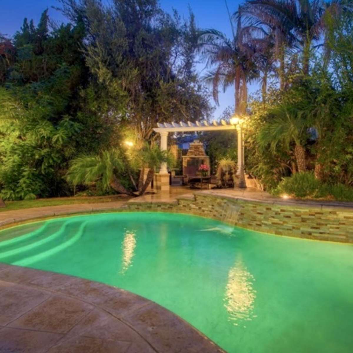 A well-lit backyard featuring a swimming pool surrounded by lush greenery and a seating area under a pergola.