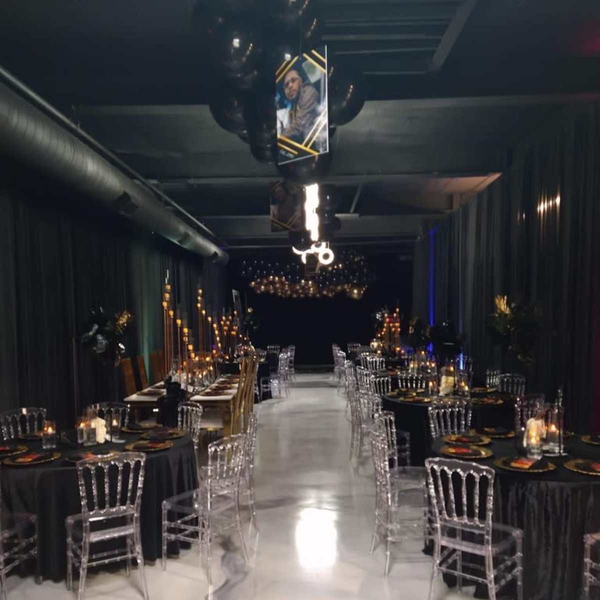 A dimly lit event space with two rows of elegantly decorated tables set for a formal dinner, featuring black tablecloths and transparent chairs.