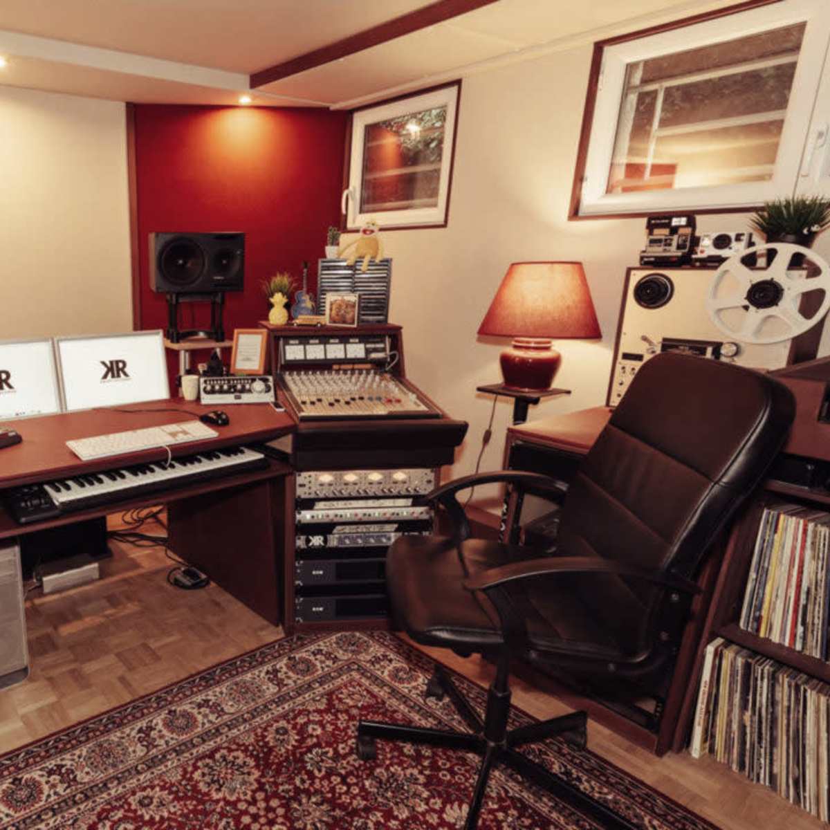 A music studio with a desk equipped with audio equipment, two computer monitors, a chair, and various recording devices against a backdrop of red and neutral walls.