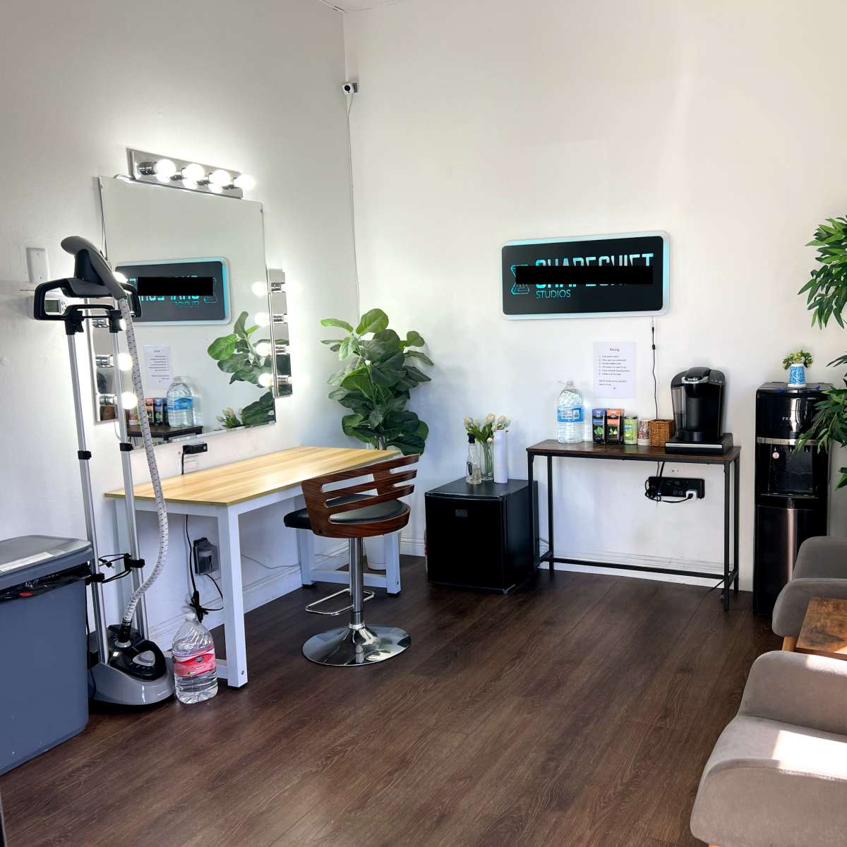 A well-lit room featuring a mirror with lights, a wooden table, a coffee station, and seating areas with plants.