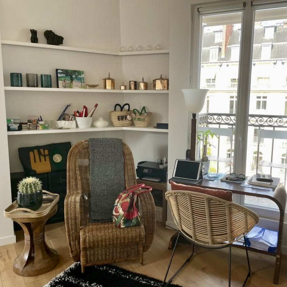 A cozy corner of a room featuring a wicker chair and small table, with shelves displaying various items and a view of a city street through large windows.