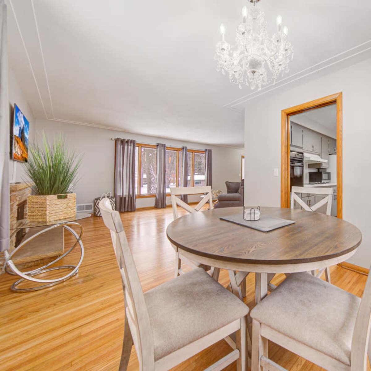 A round wooden dining table with four chairs is positioned in a well-lit dining area featuring a chandelier and an open doorway leading to a kitchen.