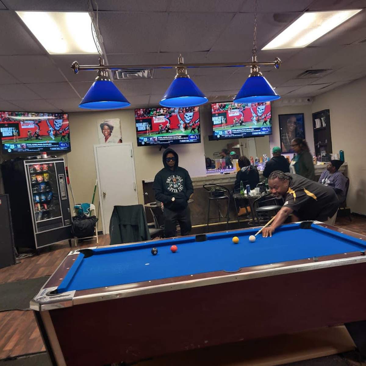 A game of pool is in progress in a dimly lit room with multiple televisions showing sports broadcasts in the background.
