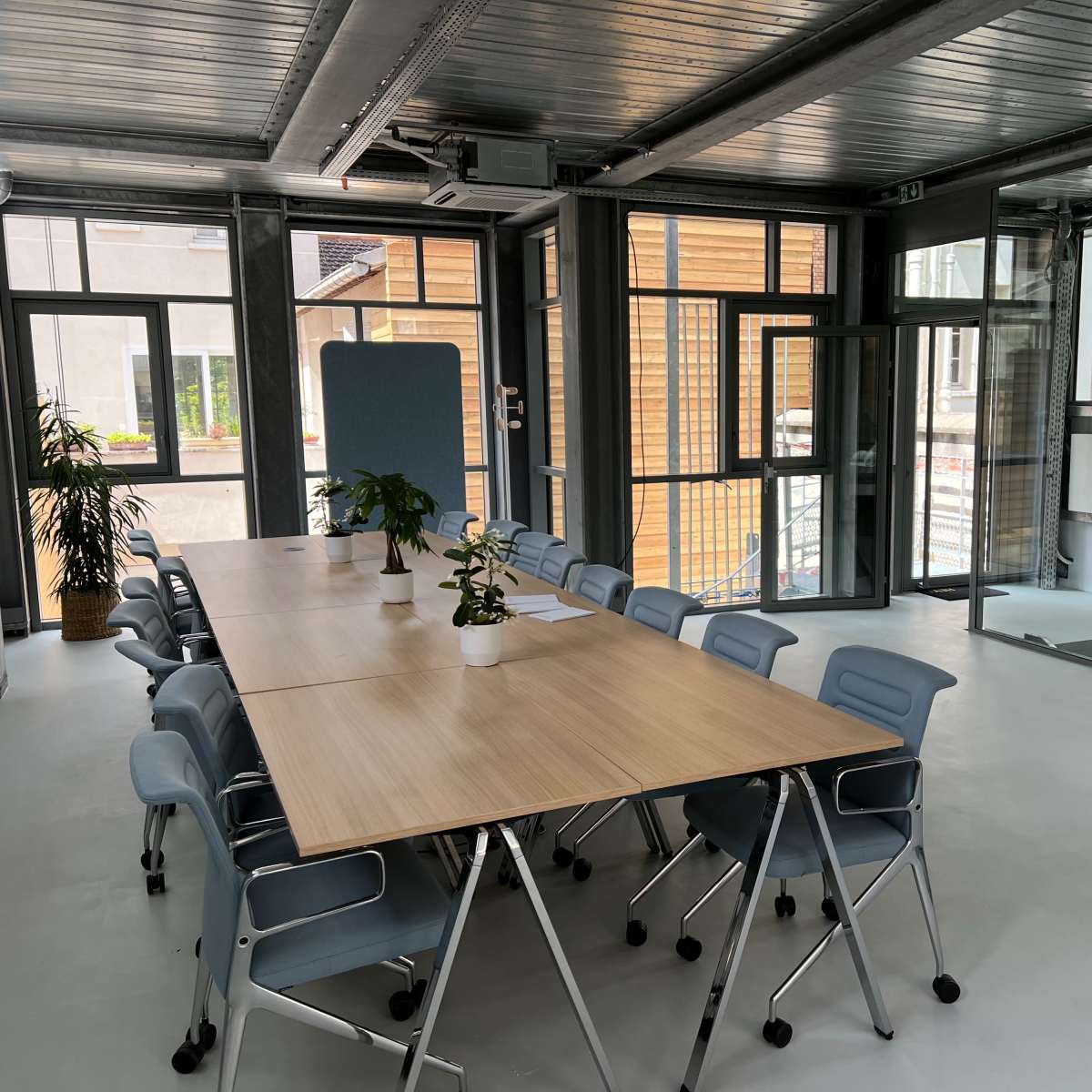A modern conference room features a long wooden table surrounded by light blue chairs, with large windows providing natural light.