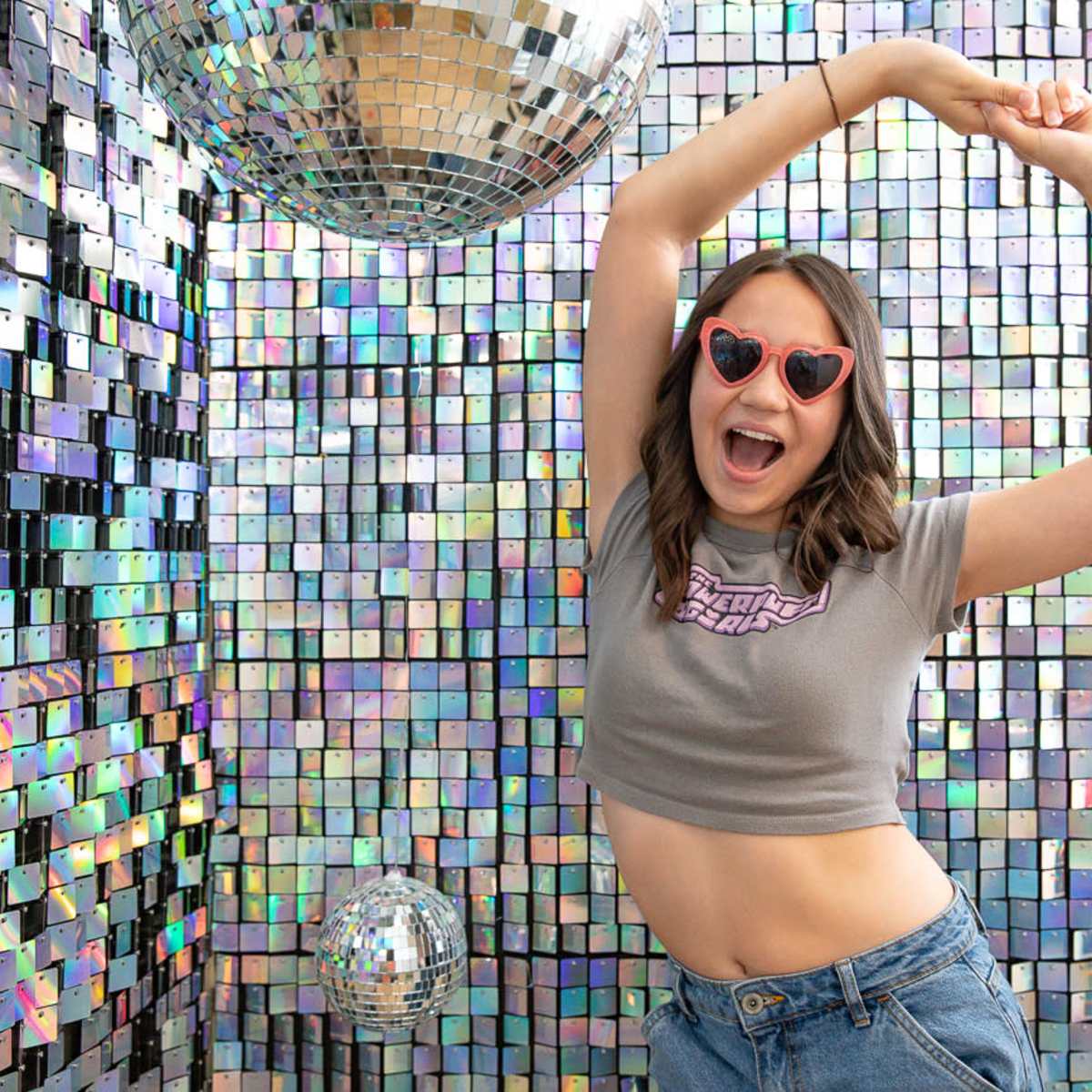A woman in a cropped shirt dances playfully in front of a shimmering, reflective wall adorned with disco balls.