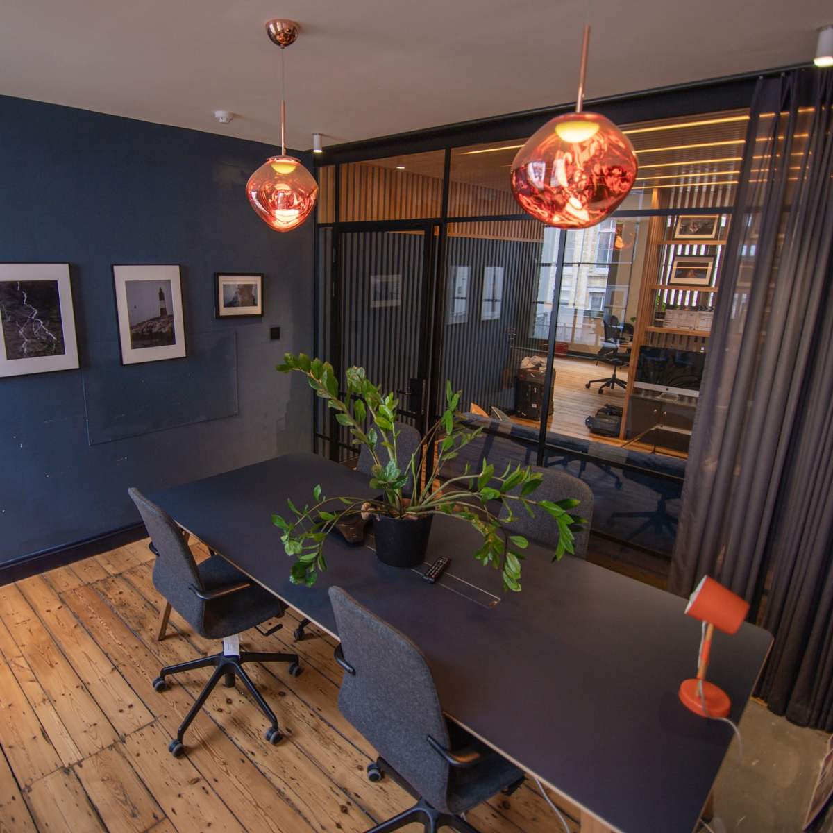 A modern office space features a long table with four chairs, a large plant centerpiece, framed photographs on the walls, and pendant lights hanging from the ceiling.