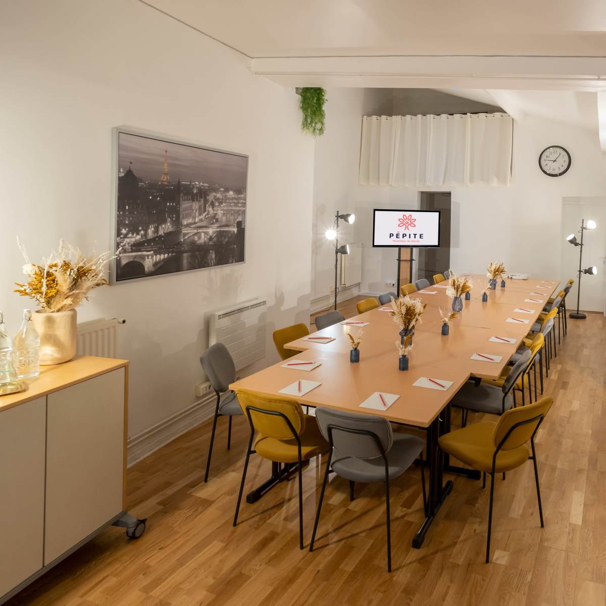 A long wooden table set for a meeting in a well-lit conference room with a wall clock, modern lamps, and decor.