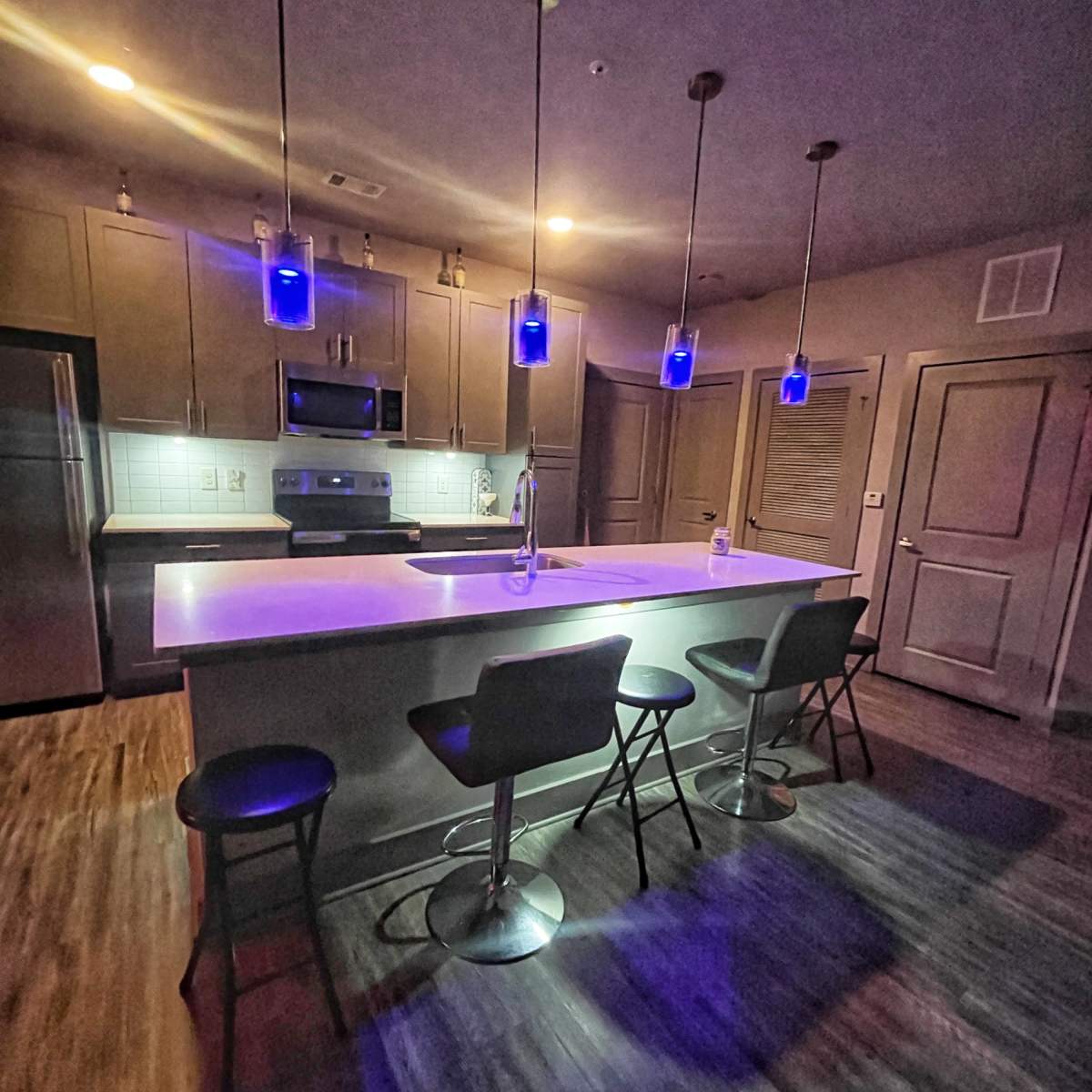 The kitchen features modern appliances, a central island with four high stools, and pendant lights illuminating the space with a blue glow.