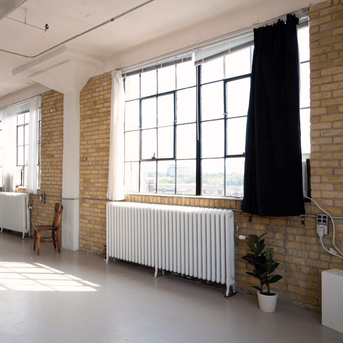 A bright, spacious room with large windows, exposed brick walls, and a few pieces of simple furniture.