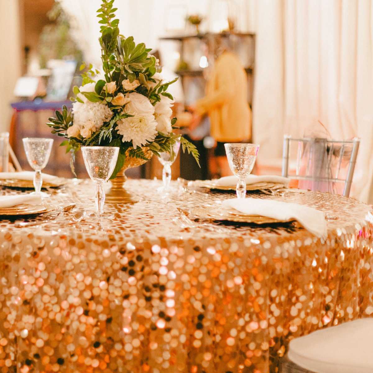 A round table covered with a gold sequin tablecloth is set for a formal meal, featuring elegant glassware and a floral centerpiece.