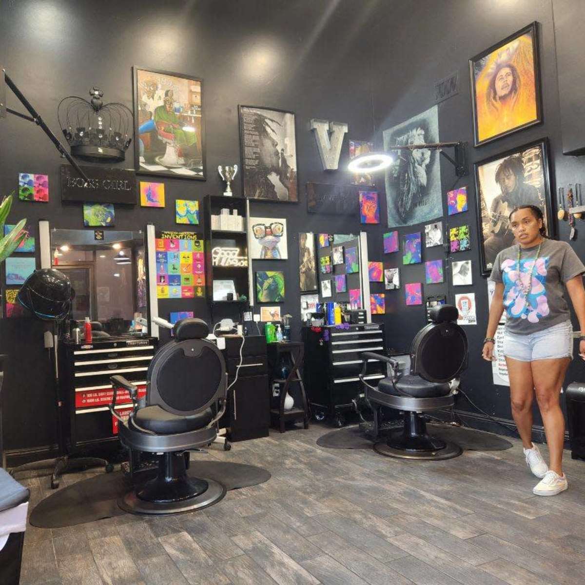 The interior of a hair salon features two black styling chairs, a wall adorned with colorful artwork, and a woman walking through the space.