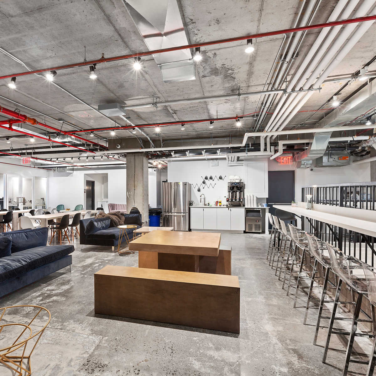 A modern office space featuring various seating areas, including a lounge with sofas, tables, and a kitchenette area with appliances.