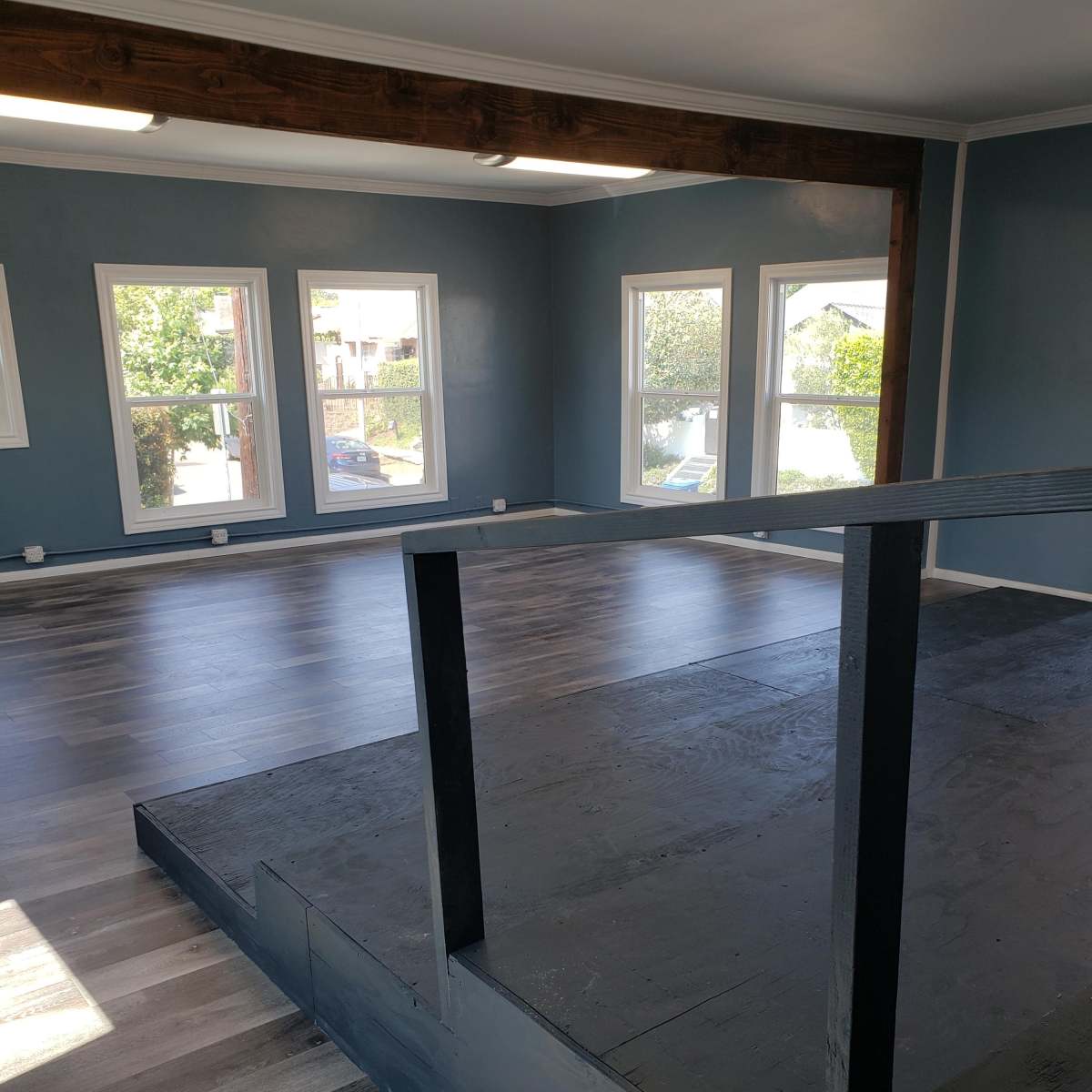 An empty room with a wooden beam across the ceiling, large windows allowing natural light, and a raised platform on the floor.