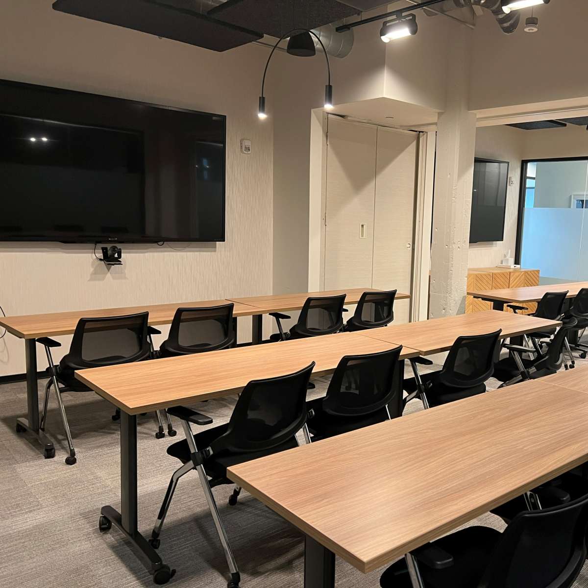 A modern conference room features multiple rows of wooden tables and black chairs, with large screens on the walls.