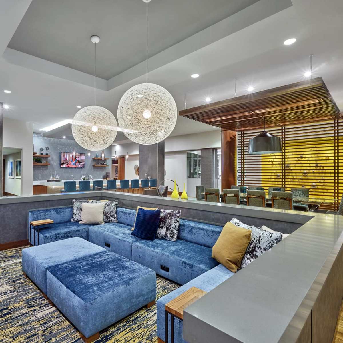 A modern living space features a blue sectional sofa with decorative pillows, two large round pendant lights, and a dining area in the background with wooden accents.