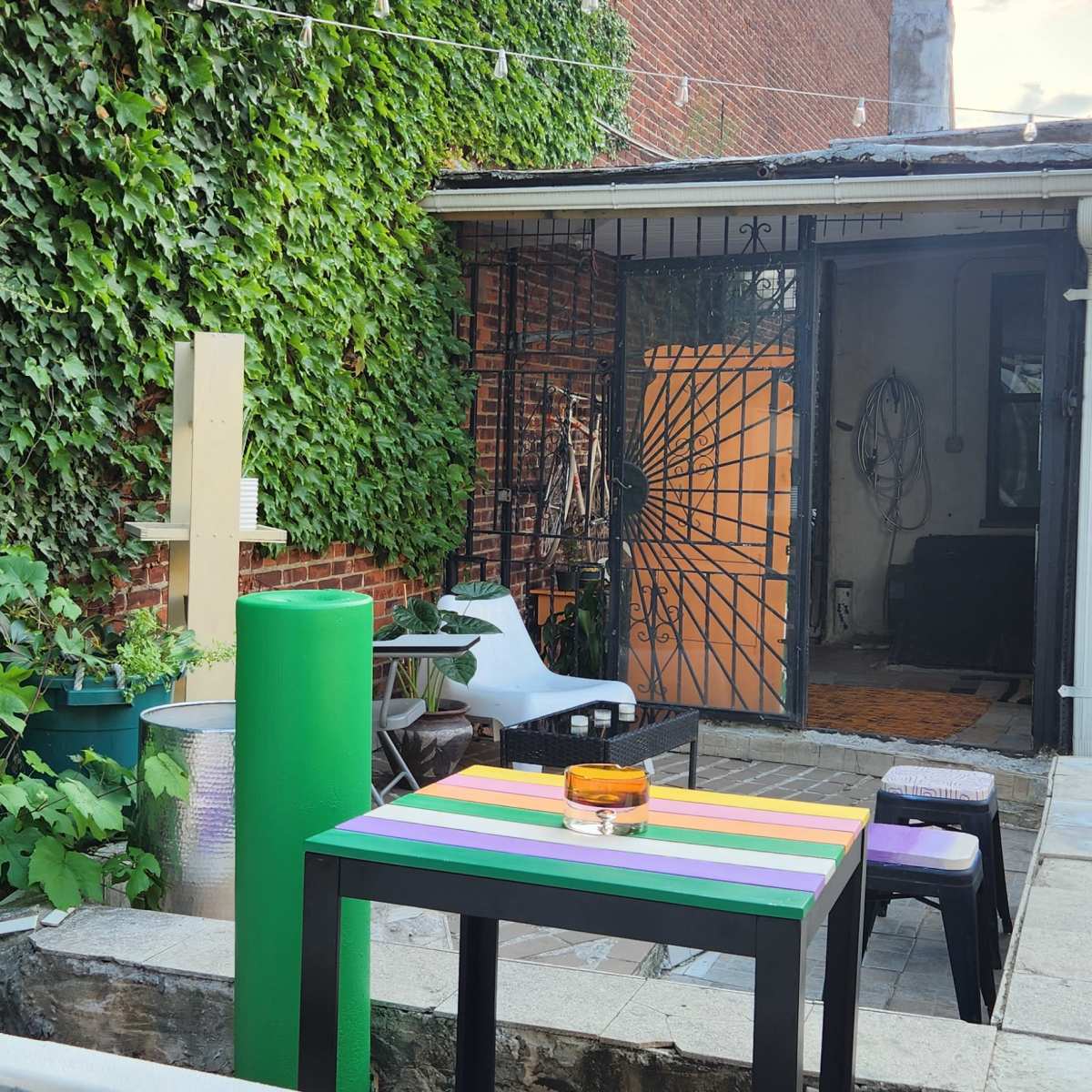 A small outdoor area with a multi-colored table, various plants, and a building with a gated entrance.