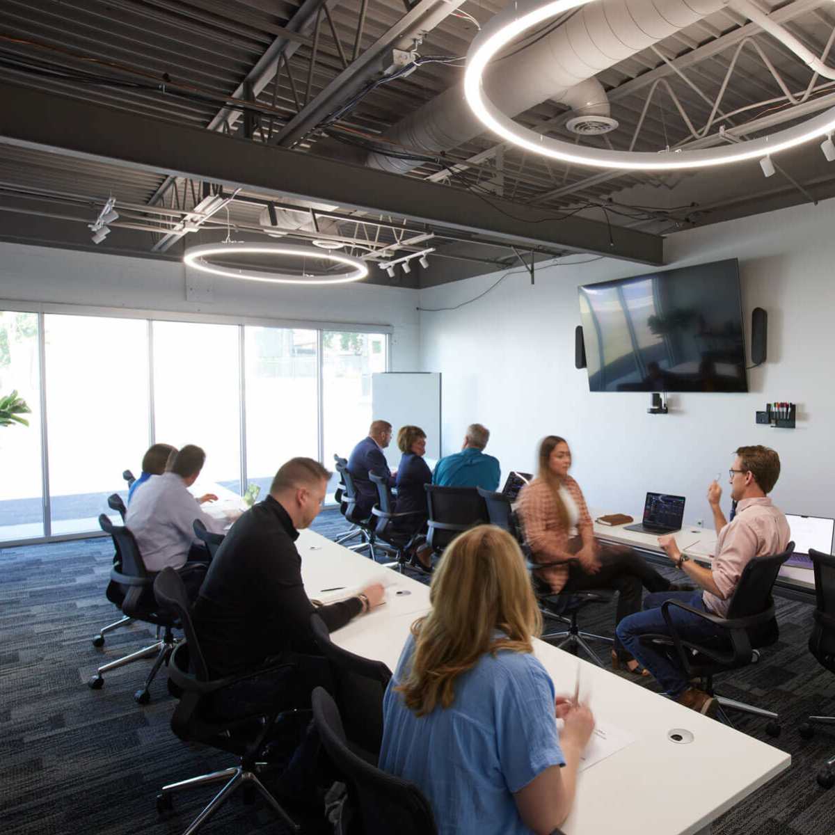 A group of people is seated at tables in a modern conference room, engaging in discussions while a presentation takes place on a screen.