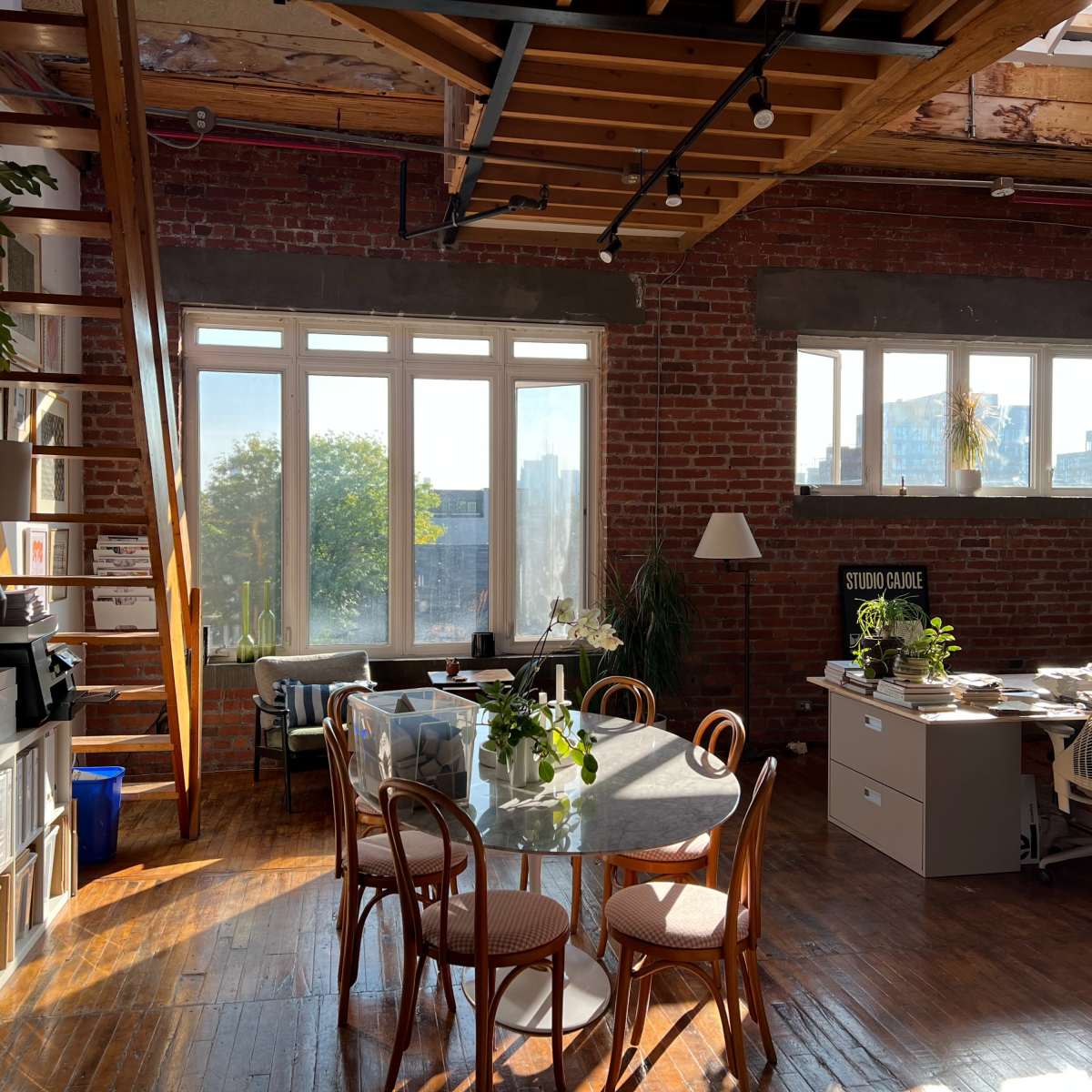 A spacious office with a round table, chairs, and natural light streaming in through large windows, featuring exposed brick walls and wooden beams.