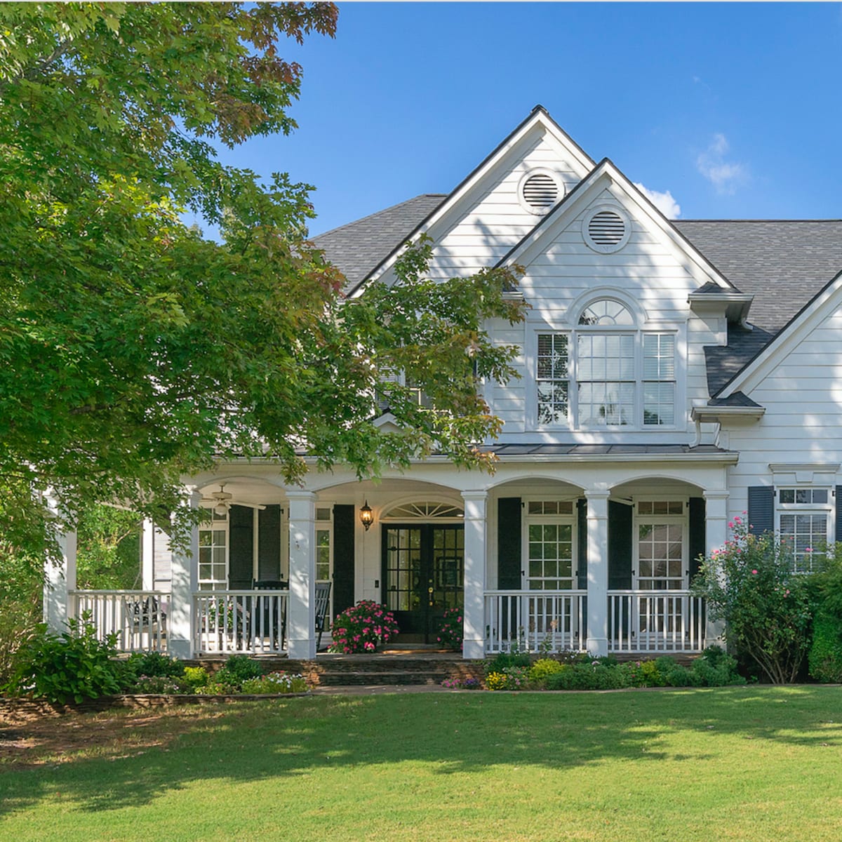 A two-story white house with black shutters and a front porch is surrounded by lush greenery and trees.
