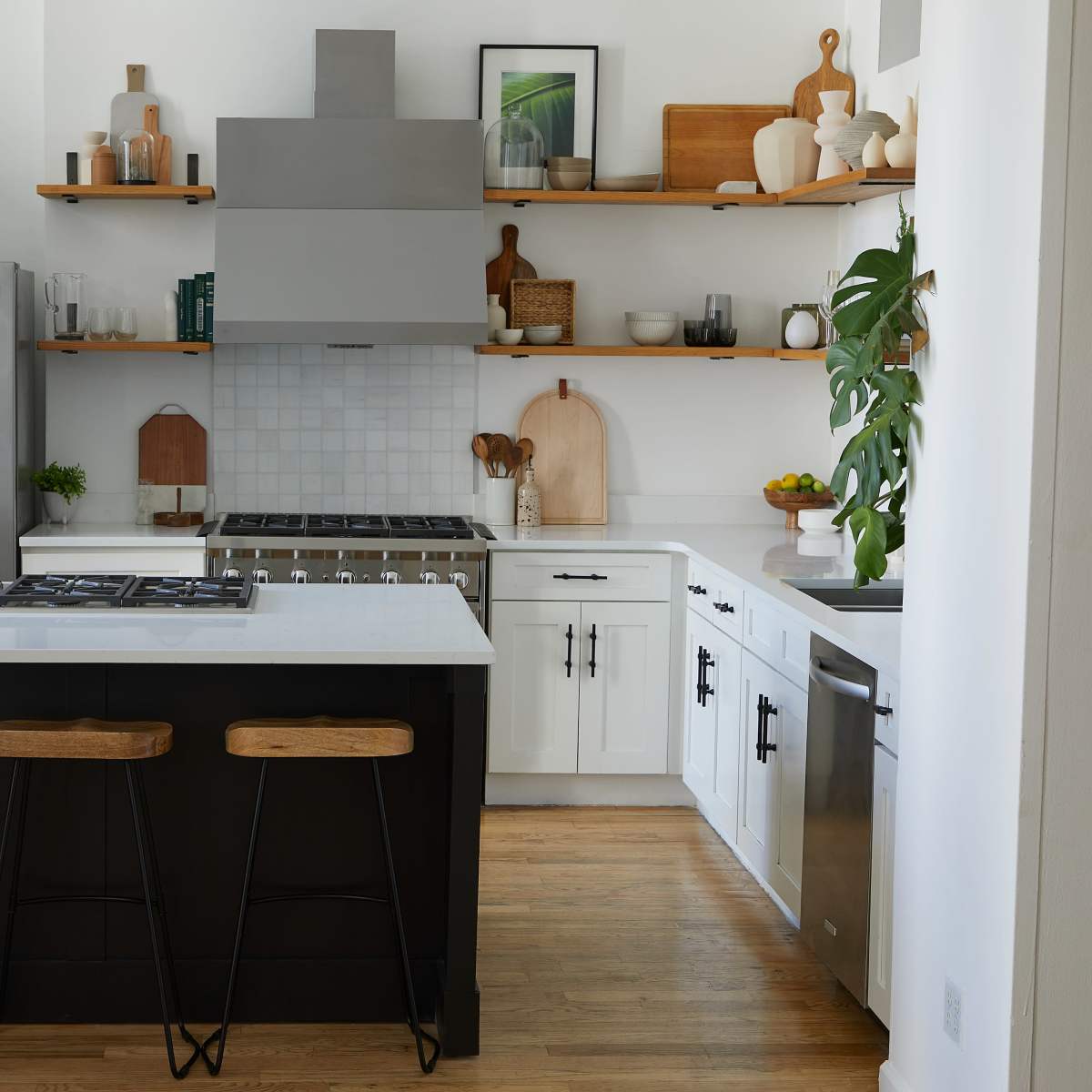 A modern kitchen features a large island with three wooden stools, stainless steel appliances, white cabinets, and open shelving, all set against a light-colored wall.