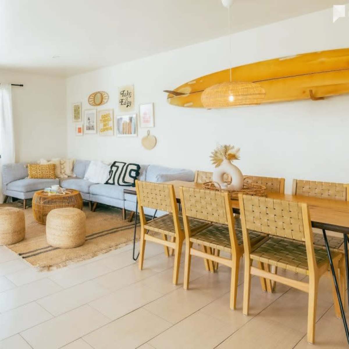 A modern living room features a light blue sofa, a wooden dining table surrounded by chairs, and decorative surfboards mounted on the wall.