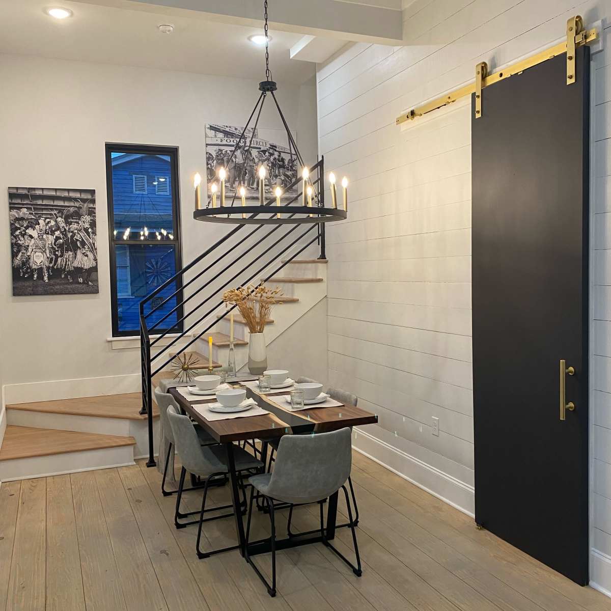 A modern dining area features a wooden table set for dinner under a chandelier, with a sliding door and staircase in the background.