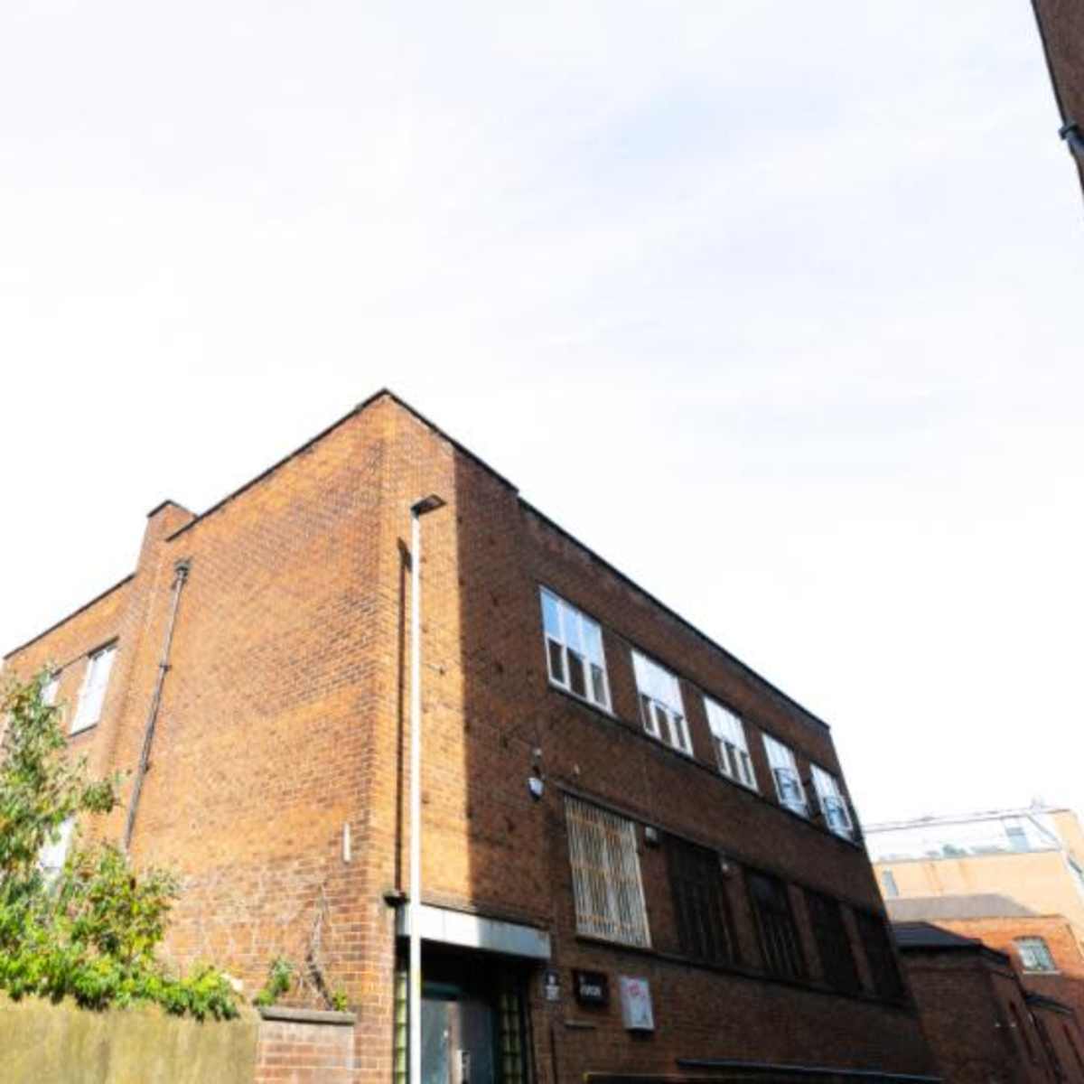 A brick building with a flat roof is seen from a low angle, surrounded by greenery and other structures.