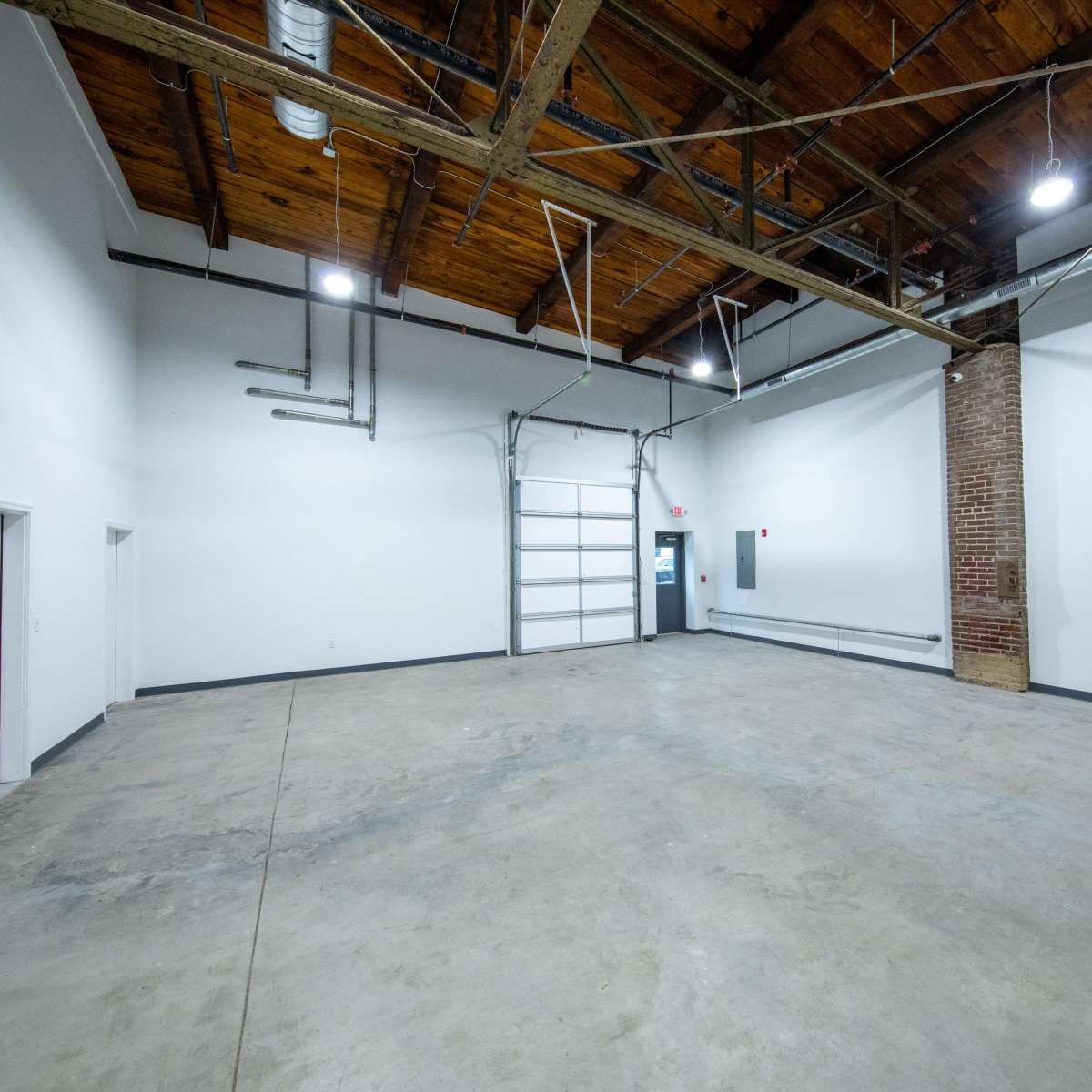 An empty industrial room with exposed wooden beams, concrete flooring, and a single door leading to another space.