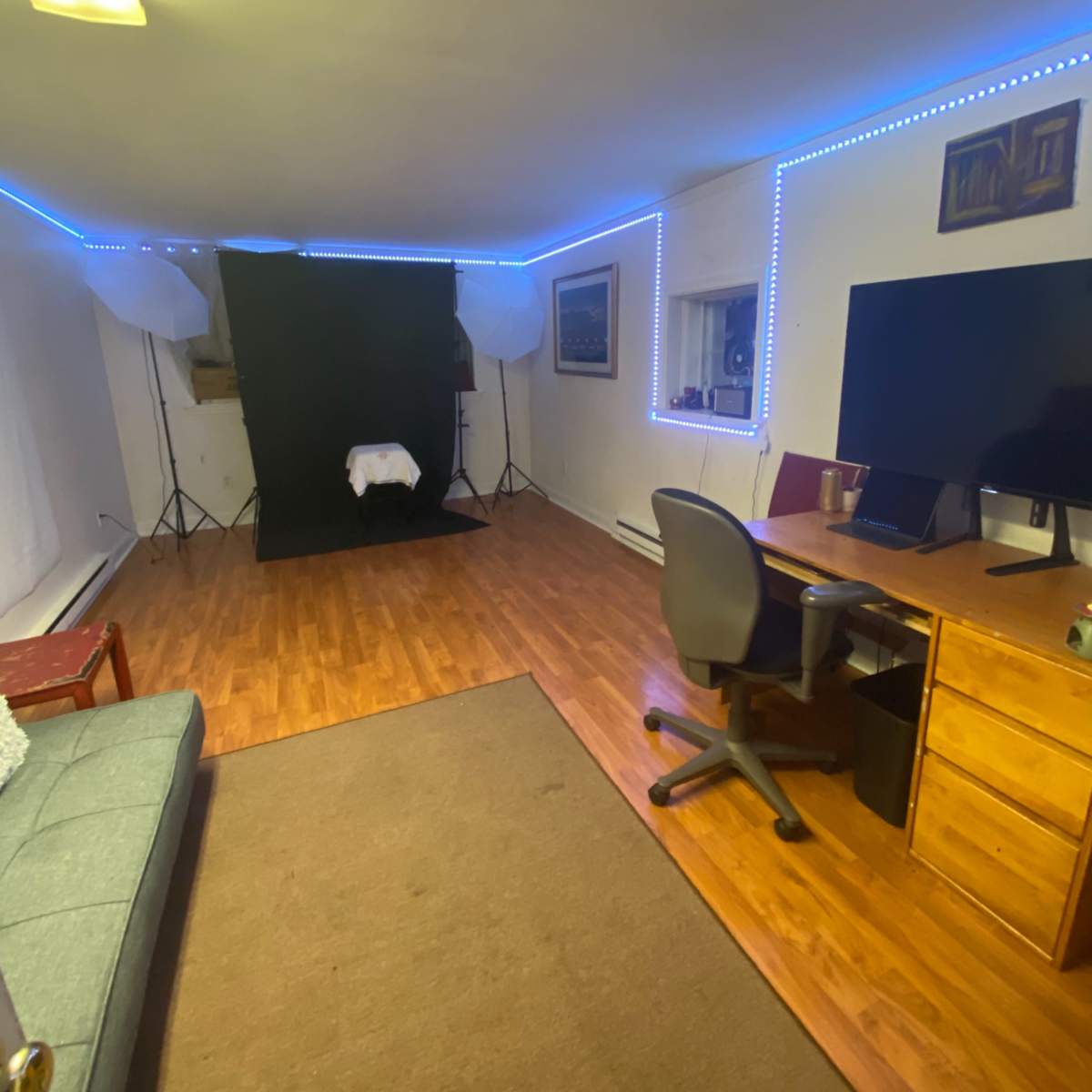 A well-lit room with wooden flooring, featuring a sofa, a desk with a computer, and a backdrop setup for photography.