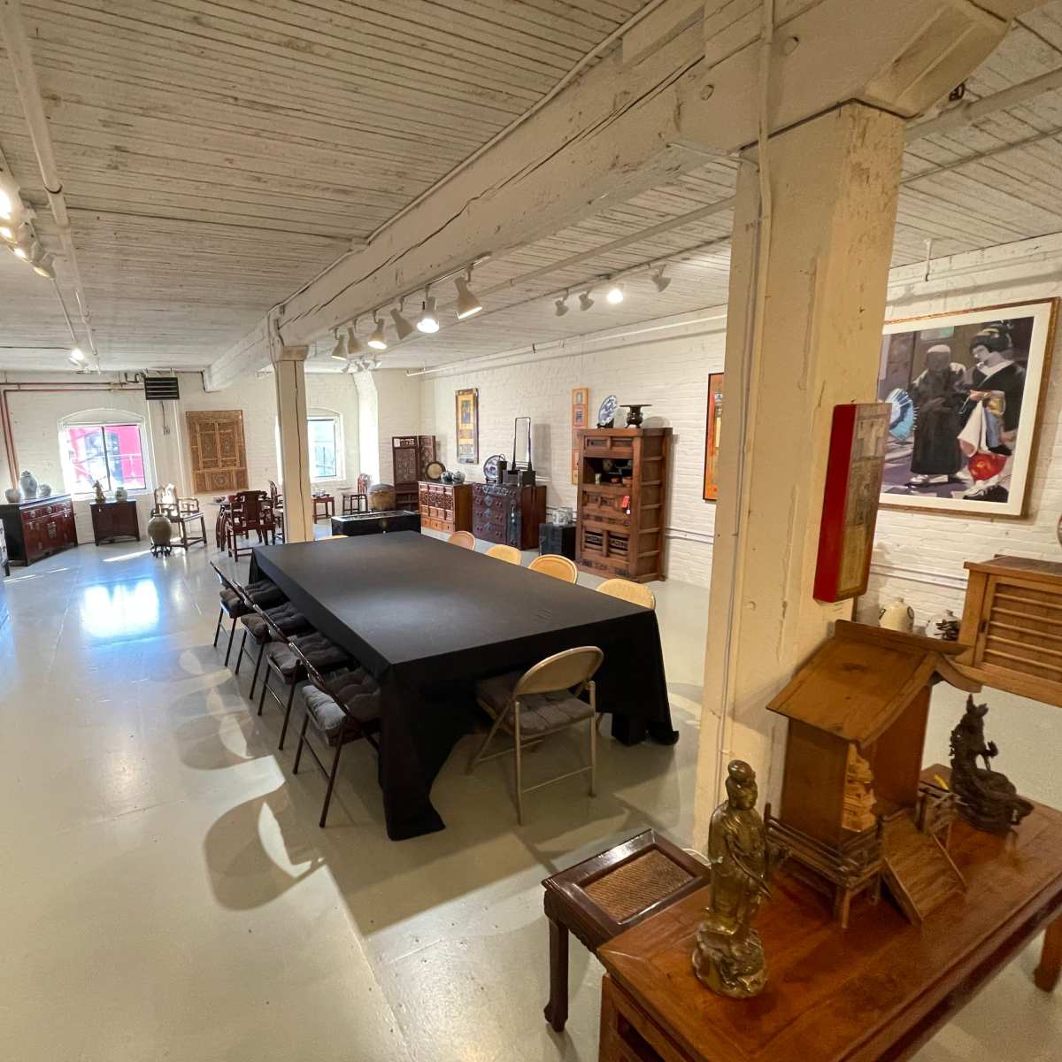 A spacious gallery room with a large black table surrounded by chairs, featuring various wooden furniture and artwork displayed on the walls.
