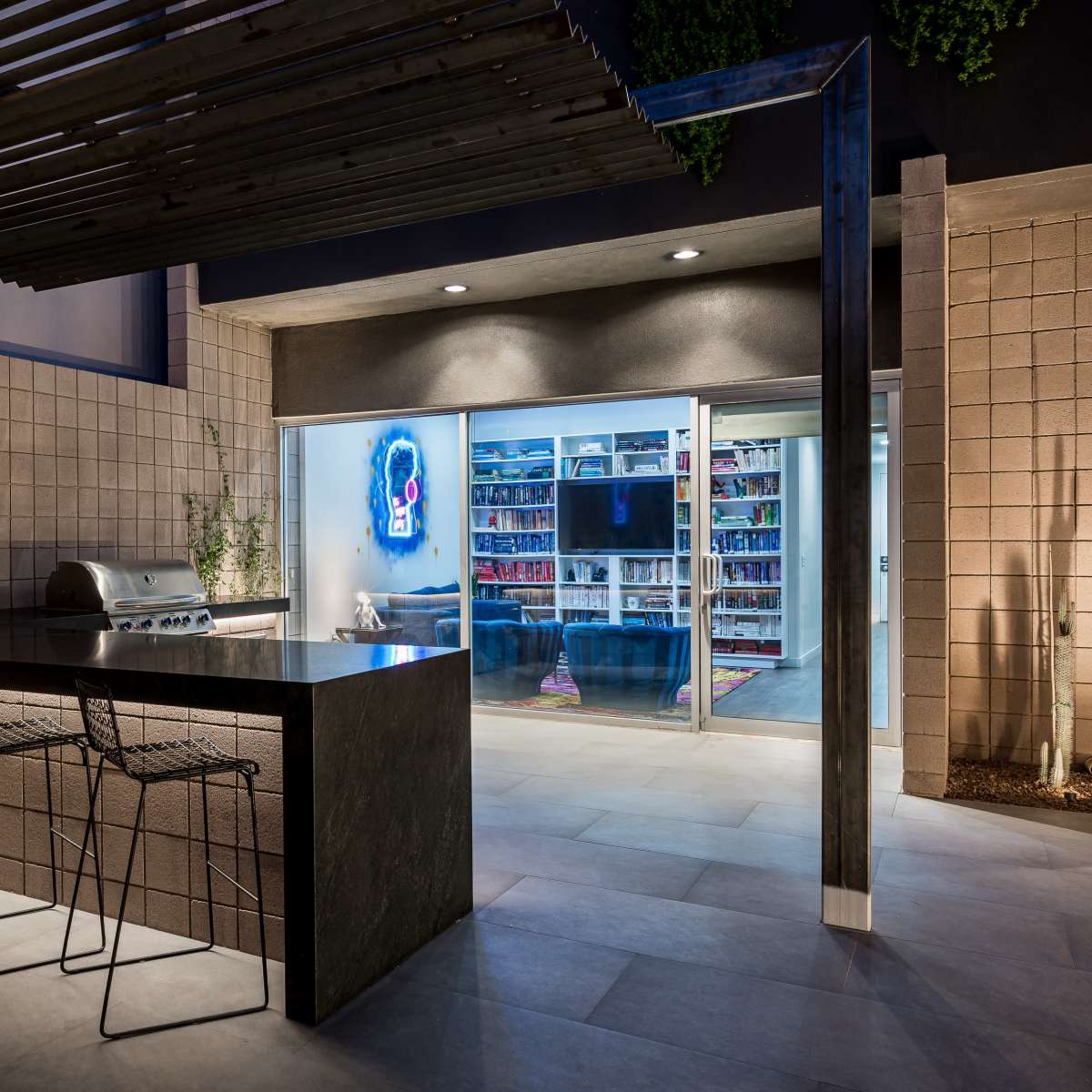 A modern outdoor kitchen with a large countertop, bar stools, and a glass door leading to a room filled with shelves of books.