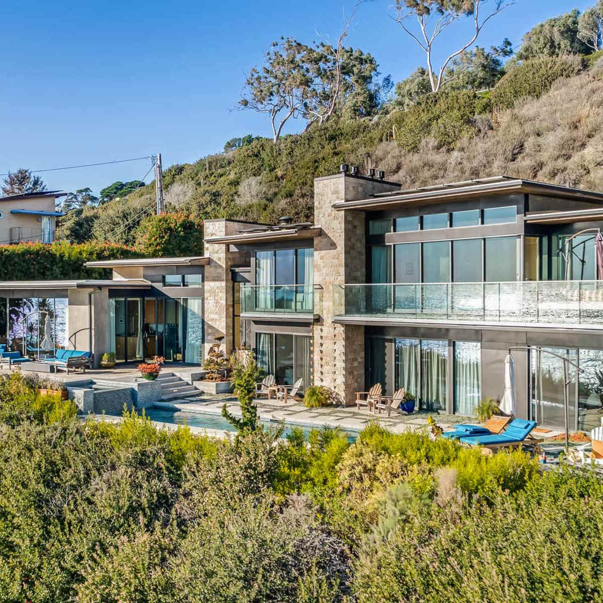 A modern hillside house with large glass windows surrounded by greenery and outdoor seating areas.